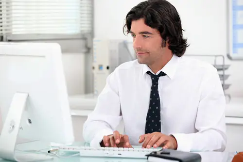 administrative worker in a shirt and tie tapping away at keyboard in front of his desktop, and there is office furniture behind him