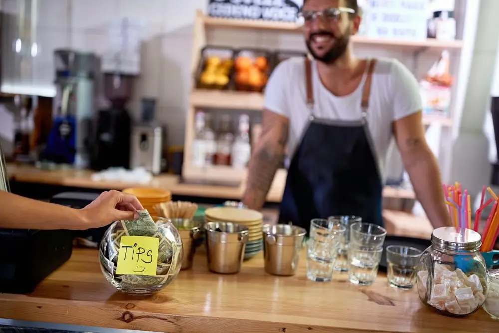 Customer at California shop putting money into a tip jar, which will be distributed to employees through a tip pool, and the server behind the counter smiles at the customer