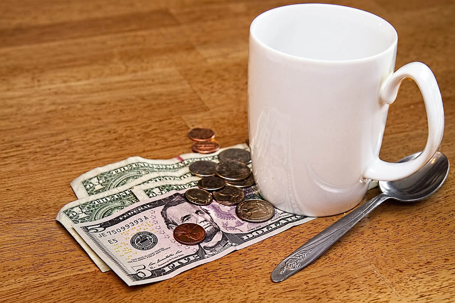 White coffee cup, a silver spoon, and about $4.50 in cash and change on a wooden restaurant table. 