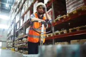 A tired warehouse worker in an orange vest and hard hat taking a momentary break.