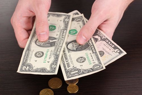 Closeup of hands holding a five dollar bill, a two dollar bill, and a one dollar bill, and underneath on the table are an assortment of coins