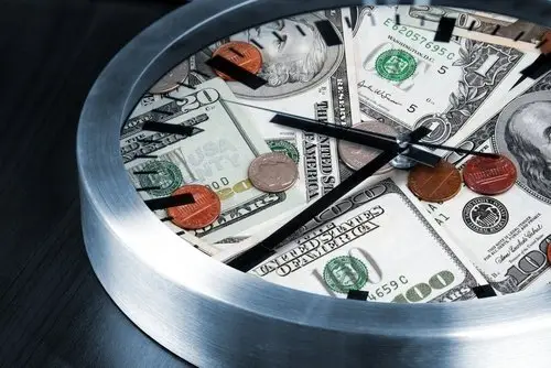 A silver clock, and the face is filled with cash and coins. The clock is laying flat in a black table. 