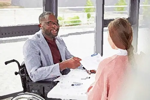 employee in a wheelchair in an office as an example of a workplace reasonable accommodation in California, and he is talking to a colleague across his desk with a large window in the backgroujd