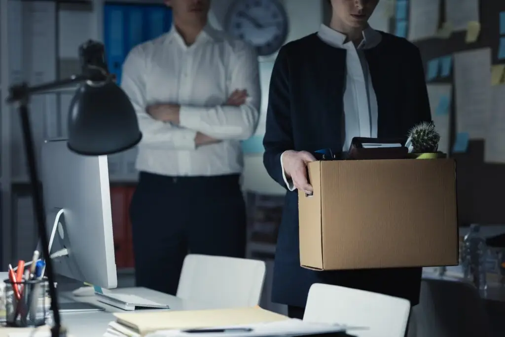 A fired employee walking out of her office carrying a cardboard box with her belongings, and her cross-armed boss is behind her watching her leave