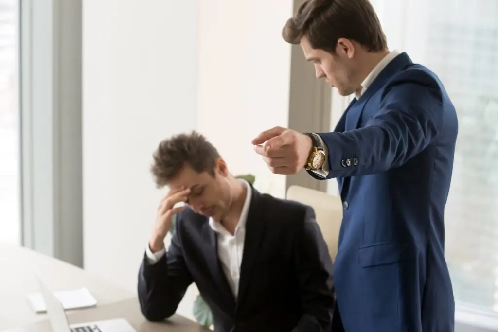 Boss in a business suit in an office, and he is berating a seated employee and pointing him towards the door.