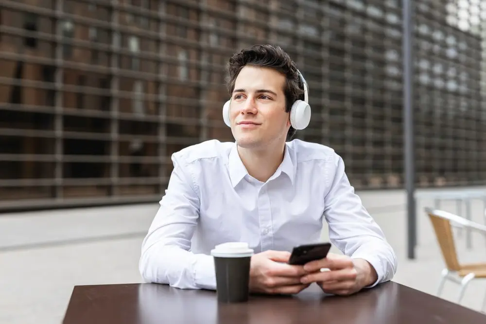 An office worker sitting outside during his break listening to music on a headset, holding his phone, and having a coffee.