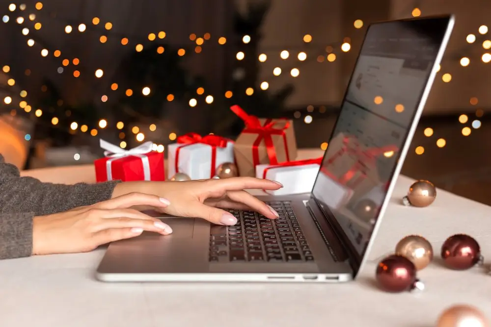 An employee working on her laptop during the holidays. In the background are holiday lights and wrapped presents, and her desk is covered in stray ornaments. 